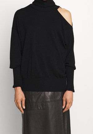 Pull en maille noire avec col haut, coupe oversize et une épaule dénudée. Poignets et ourlet côtelés ; porté avec une jupe en cuir marron.