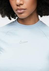Jasnoniebieski sportowy top z okrągłym dekoltem i krótkimi rękawami. Z przodu znajduje się srebrne logo Nike, a materiał jest gładki i elastyczny.