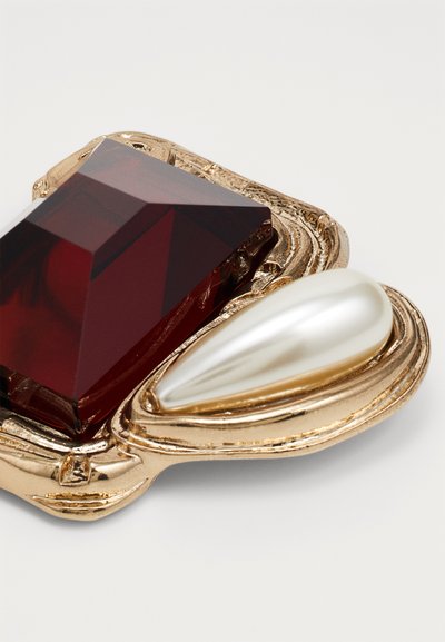 Gold-tone brooch featuring a large red gem with a faceted shape and a teardrop-shaped white pearl accent. Textured, wavy gold setting.