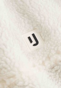 Vitt, fluffigt fleece-material med en texturerad yta. Har en fyrkantig vit etikett med en svart stiliserad "J"-logotyp.