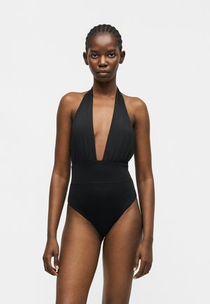 Γυναίκα με πλεξούδες που φοράει μαύρο ολόσωμο μαγιό με τιράντα γύρω από το λαιμό και βαθύ V ντεκολτέ, στέκεται μπροστά σε απλό φωτεινό φόντο.