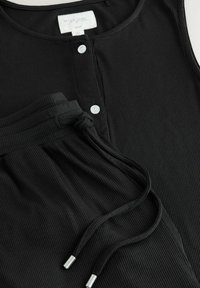 Débardeur noir à côtes avec boutons-pression blancs et pantalon à cordon assorti. Texture douce avec une coupe ajustée et des accents argentés sur le cordon.