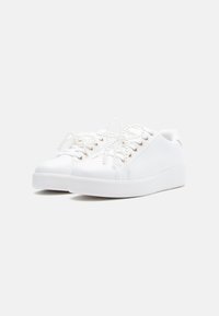 Baskets synthétiques blanches avec semelles en caoutchouc plates, dotées de lacets ronds, d'œillets, et d'un design minimaliste. Texture lisse et style bas.