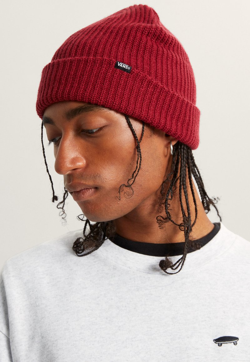 Red ribbed knit beanie with a pointed top and small black Vans logo tag. Model wears a light gray sweatshirt with dark neckline.