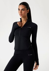 Black athletic jacket with zipper, long sleeves, and fitted design. Features "GUESS" logo on chest, paired with matching black leggings.