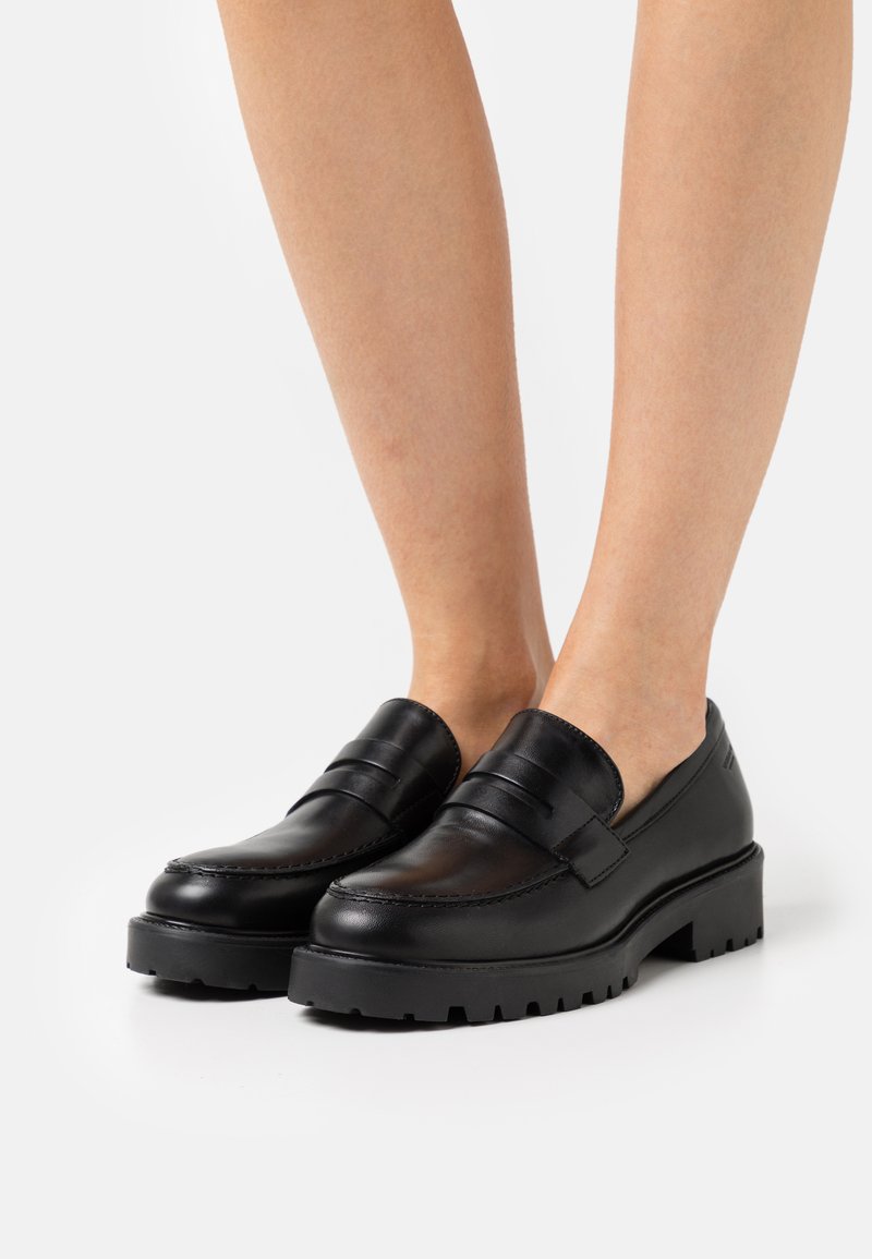 Mocassins de couro negro com sola de tração grossa usados em pés descalços, mostrando a vista frontal e lateral contra um fundo branco.