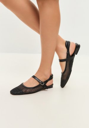 Picioare purtând balerini slingback negri din plasă cu o curea cu cataramă peste podul piciorului, prezentate pe un fundal deschis simplu.