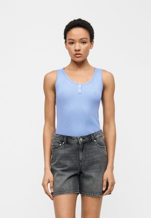 Žena stojaca čelom vpred, oblečená v svetlomodrom rebrovanom tielku bez rukávov s gombíkmi a v čiernych vyblednutých krátkych rifliach s vysokým pásom.