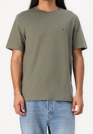 Homme portant un t-shirt vert olive à manches courtes avec un petit logo sur la poitrine et un jean bleu clair.
