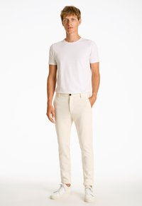 T-shirt bianco in cotone abbinato a pantaloni crema a gamba affusolata e sneakers bianche. I pantaloni presentano una zip frontale e un singolo dettaglio con bottone nero.