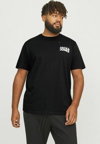 Schwarzes Baumwoll-T-Shirt mit kurzen Ärmeln, das ein weißes Logo auf der linken Brust zeigt. Der Stoff wirkt glatt und leicht.