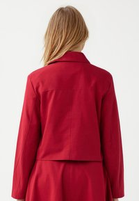Czerwona dopasowana marynarka z wykładanym kołnierzem, długimi rękawami i skróconym tyłem. Wykonana z gładkiego materiału z minimalnymi detalami przeszycia.