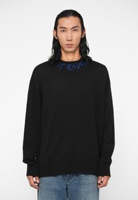 Črna pulover s svobodnim krojem in rebrastimi manšeti. Ima modro vezeno besedilo ob izrezu. Obučen je z svetlo modrimi kavbojkami.