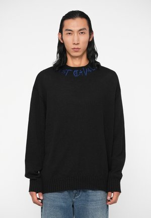 Pull noir à coupe ample avec des poignets côtelés. Présente un texte brodé en bleu le long du col. Porté avec un jean en denim bleu clair.