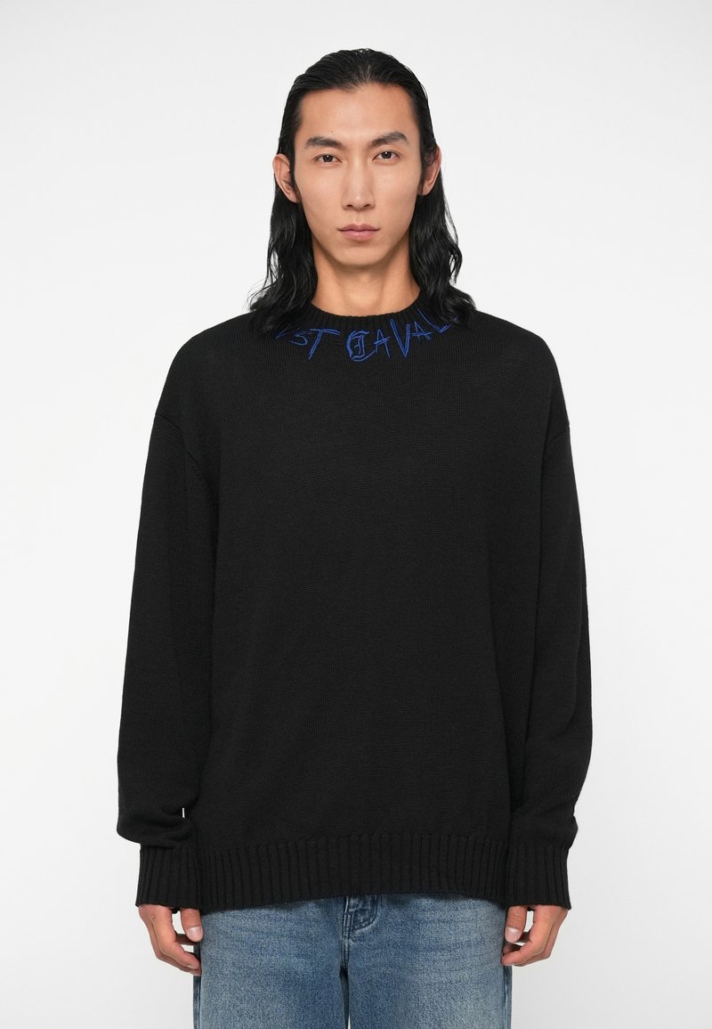 Črna pulover s svobodnim krojem in rebrastimi manšeti. Ima modro vezeno besedilo ob izrezu. Obučen je z svetlo modrimi kavbojkami.