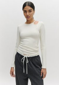 Witte lange mouwen top met ribbelstructuur en ronde halslijn, met een uitsnijding op één schouder en knoopaccenten. Gecombineerd met donkergrijze broek.