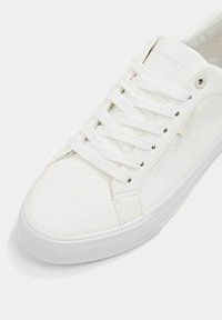 Weißer Sneaker mit glattem synthetischem Obermaterial, flachen weißen Schnürsenkeln, Gummisohle und einem dezenten Logodetail an der Seite.