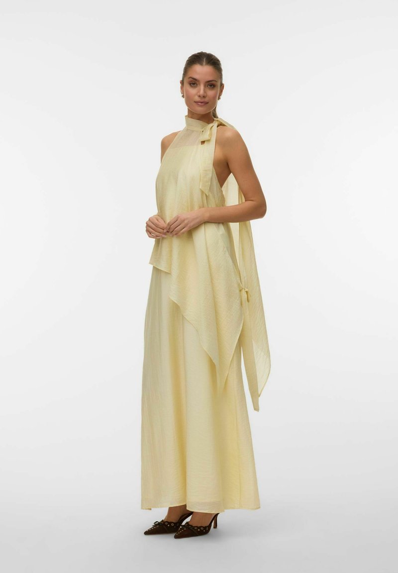 Femme portant une longue robe maxi jaune pâle à col halter avec un tissu drapé asymétrique et des talons pointus foncés, debout devant un fond blanc.