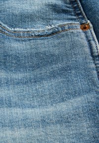 Šviesiai mėlynos spalvos džinsinio audinio, šiek tiek nudėvėta kišenės sritis, su geltonais siūlais ir mažu rudos spalvos metaliniu vinuku.