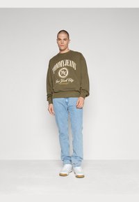 Felpa verde oliva con logo "TOMMY JEANS" e jeans azzurri chiari. Chi la indossa ha anche sneakers bianche con suola in gomma e uno sfondo neutro.