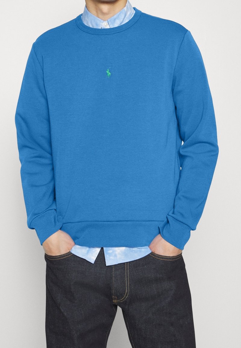 Personne portant un sweat bleu à col rond sur une chemise à col bleu clair avec un jean foncé, les mains dans les poches, face à l'avant.