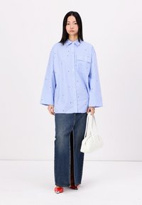 Chemise oversize rayée bleu et blanc avec une poche avant, associée à une jupe en jean foncé taille haute et un sac à main blanc. Chaussures rouges visibles.