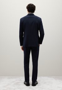 Hombre de pie frente a una pared, vistiendo un traje a rayas azul marino y zapatos de vestir negros sobre un suelo alfombrado de color gris claro.
