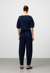 Vrouw met een knot in haar haar, gekleed in een marineblauwe losse top met ellebooglange mouwen en bijpassende hoge broek, staande op een grijze vloer.
