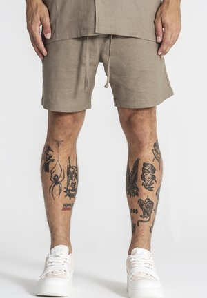 Man die beige korte broek en witte sneakers draagt, toont getatoeëerde benen met verschillende zwarte inktontwerpen, waaronder een spin, een hond, een adelaar en een vrouwen gezicht.