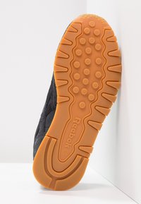 Svart sneaker med en texturerad ovandel och gummisula. Sulan är orange med cirkulära grepp och ett zigzag-mönster för bättre fäste.