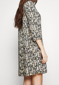 Robe fleurie avec un fond noir, ornée de fleurs blanches et jaunes, manches trois-quarts, design plissé, et tissu léger.