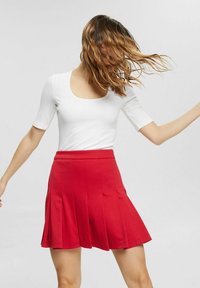 Haut blanc ajusté avec un col rond et des manches courtes, associé à une jupe plissée rouge qui s'évasent au niveau de l'ourlet. Texture de tissu lisse.