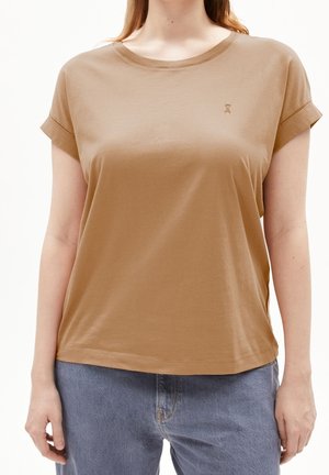 Frau trägt ein hellbraunes T-Shirt mit kurzen Ärmeln und aufgerollten Bündchen sowie blaue Jeans, steht vor einem weißen Hintergrund.