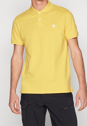 Muž v žluté krátkosleevé polokošili s bílým logem stromu na hrudi a černých kalhotách typu cargo.