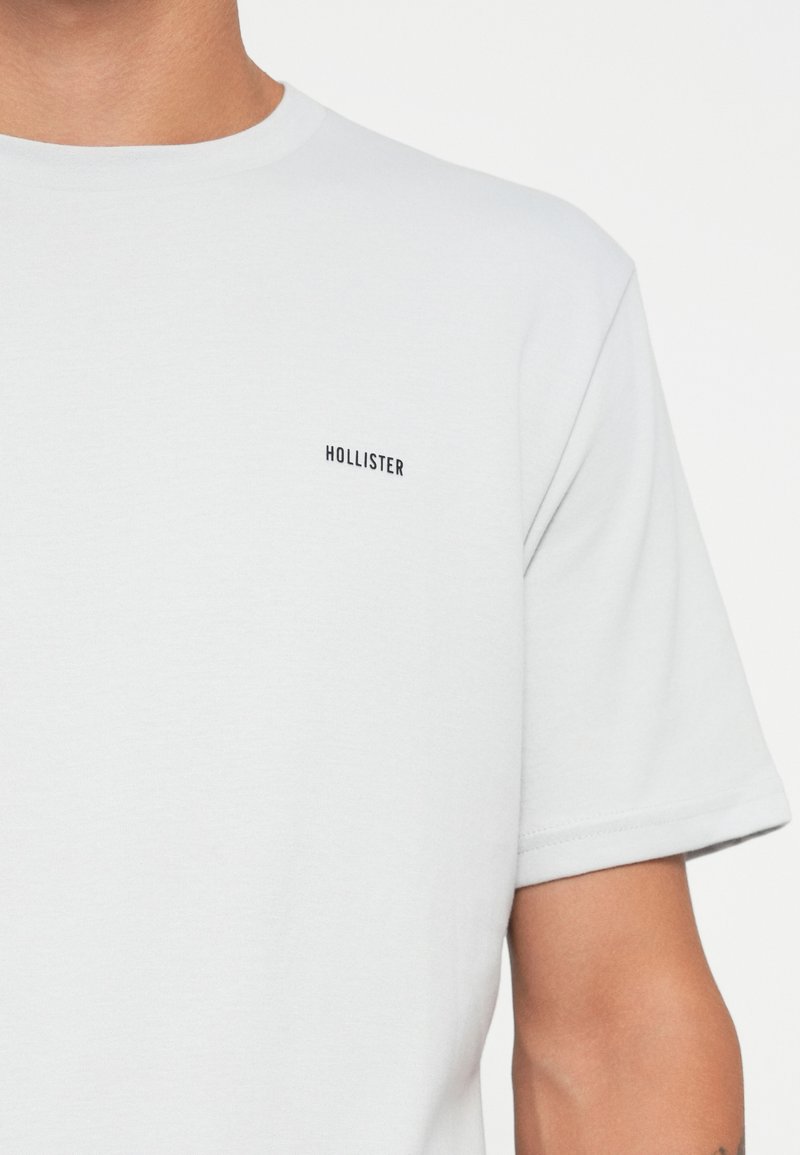 Camiseta blanca de manga corta hecha de algodón. Presenta un pequeño logo negro "HOLLISTER" en el lado izquierdo del pecho. Textura suave y diseño clásico de cuello redondo.