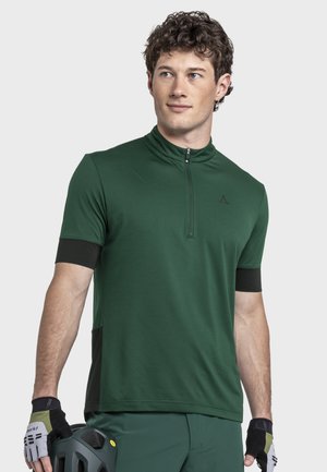 Mann trägt dunkelgrüßes kurzärmeliges Zip-up-Shirt und Handschuhe, hält schwarzen Fahrradhelm und schaut leicht nach links vor grauem Hintergrund.