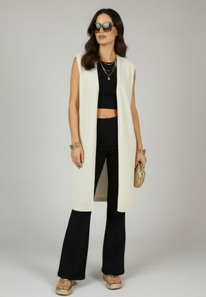 Vrouw draagt oversized zonnebril, mouwloos wit gebreid vest, zwarte crop top, zwarte uitlopende broek, gouden sandalen, armbanden, gelaagde halskettingen, houdt een gouden clutch vast.