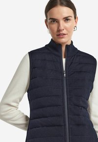 Marinblå vadderad väst med hög krage och vertikal sömnad, dragkedjestängning, vid användning över en ljusfärgad topp, med en slät tygtextur.