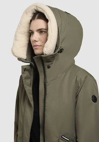 Olivi vihreä huppuparka, jossa on kermaisen värinen tekoturkisvuori, vetoketjukiinnitys ja musta logoaksentti. Ominaisuuksiin kuuluu sileä ja kestävä kangas.