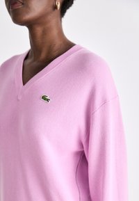 Suéter de cuello en V rosa con un tejido texturizado, presenta un pequeño logo de cocodrilo verde en el lado izquierdo del pecho. Corte relajado con hombros caídos.