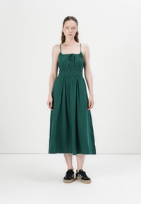 Grön, ärmlös maxiklänning med smockad byst och justerbara band. Kombineras med svarta sneakers med en ljusfärgad sula.