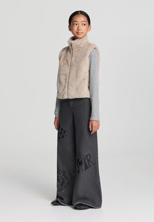 Gilet in pelliccia sintetica beige con colletto alto, maglietta grigia con maniche lunghe e jeans in denim scuro a gamba larga con stampe grafiche e orlo sfrangiato.