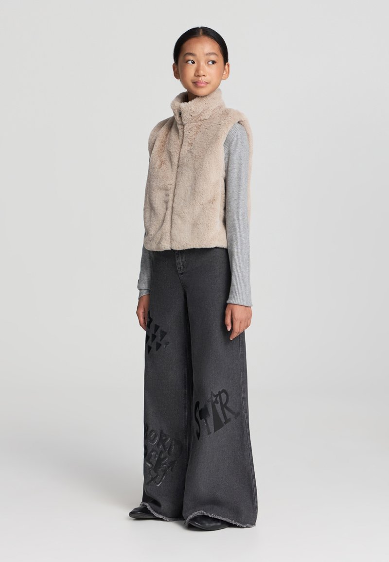 Gilet in pelliccia sintetica beige con colletto alto, maglietta grigia con maniche lunghe e jeans in denim scuro a gamba larga con stampe grafiche e orlo sfrangiato.