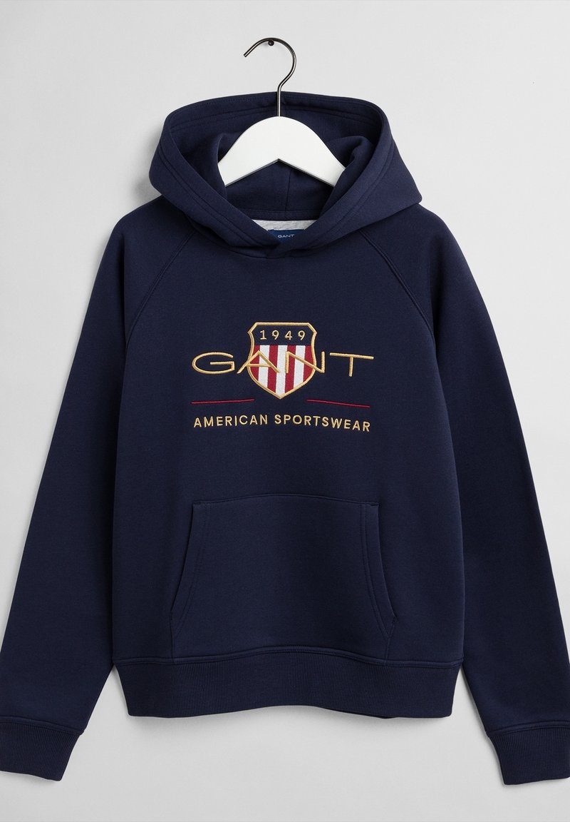 Sweat à capuche bleu marine avec poche avant, logo écusson brodé "GANT 1949" et texte "American Sportswear" sur la poitrine.