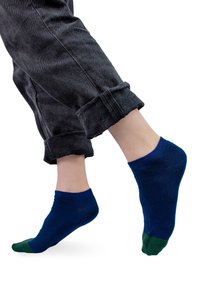 Niebieskie skarpety z zielonym palcem; miękki dzianinowy materiał. Noszone z podwiniętymi ciemnoszarymi dżinsami, podkreślającymi szczegóły kostki i skarpet.