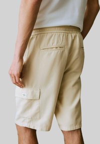 Beige shorts med elastisk midjeband, som har två bakfickor och en sidoficka för cargobyxor. Tillverkade av ett lätt och andningsbart material.