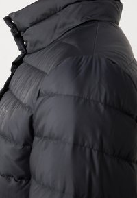 Gros plan sur l'épaule et le col montant d'une doudoune noire matelassée avec un tissu mat et des détails de coutures horizontales.