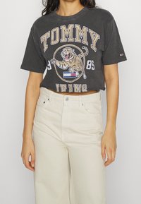 Camiseta negra corta con un gran gráfico de un león, texto en amarillo y blanco que dice "TOMMY JEANS", combinada con pantalones de tiro alto de color beige claro.