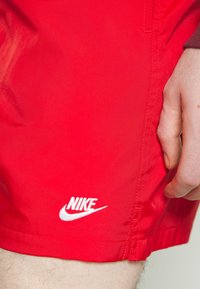 Gros plan sur une personne portant un short rouge Nike avec le logo blanc de Nike près de l'ourlet et une main reposant à côté du short.