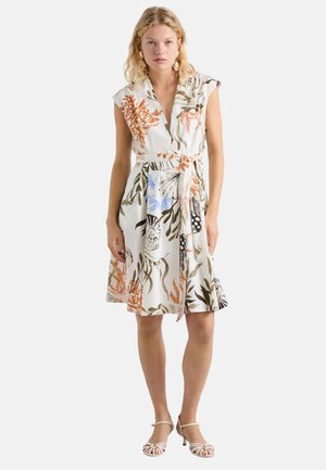 Donna che indossa un vestito bianco senza maniche, lungo fino al ginocchio, con stampe floreali e di conchiglie, annodato in vita, abbinato a tacchi beige con cinturini.